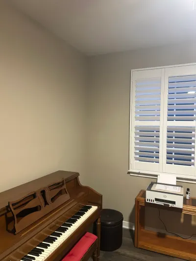 Corner room with brown upright piano, black ottoman, and window in white plantation shutters.