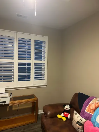 Corner of a den with white plantation shutters, greige walls, LVP flooring and a brown leather sofa.