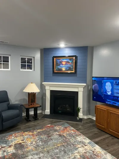 Finished basement living area with blue shiplap fireplace wall, white mantel, TV, recliner and patterned rug.