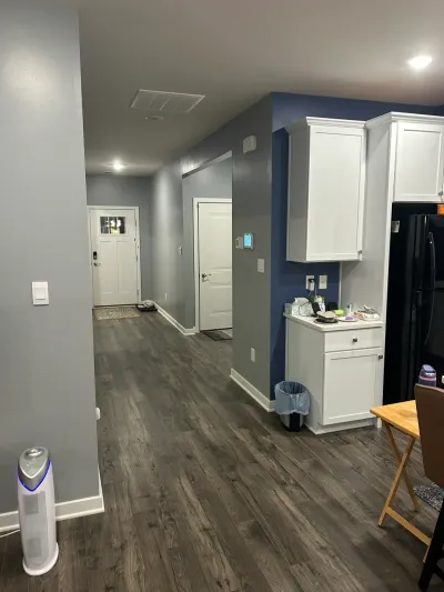 Gray hallway with wood-look vinyl plank floor leading to a white front door, beside white kitchen cabinets on a navy wall.