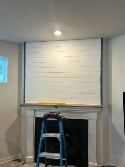 Fireplace wall mid-remodel with white shiplap, painter's tape, raw mantel shelf and a step ladder in front.