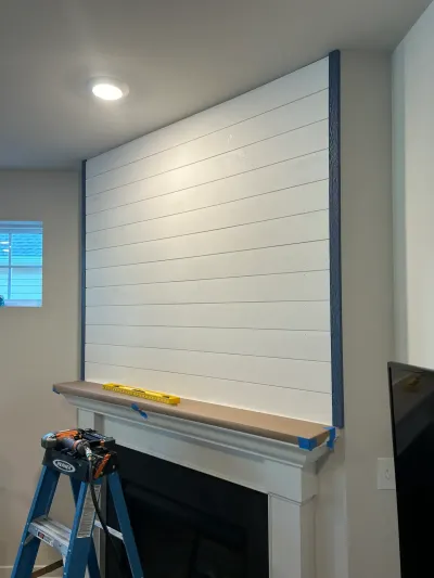 Fireplace wall mid-remodel with new white shiplap, taped mantel shelf, level, ladder and nail gun.