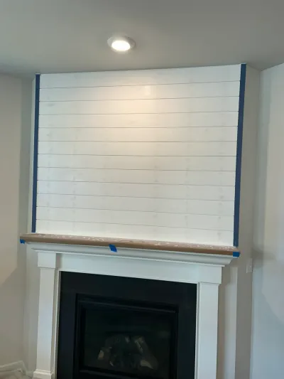 Fireplace with white shiplap surround mid-painting, blue tape at edges and paper-covered mantel.