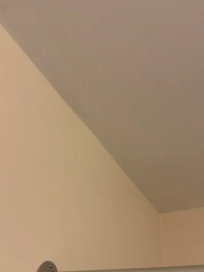 Upper corner of a room where a sloped ceiling meets cream painted walls, with a metal bracket at the bottom edge.