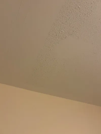 Close-up of a beige wall and ceiling corner showing a mismatched textured drywall patch repair.