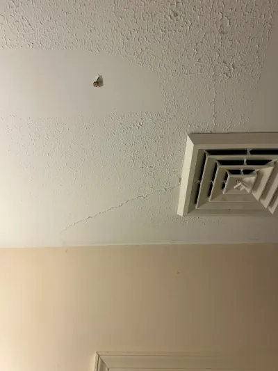 Textured white ceiling with a mismatched drywall patch, hairline crack, and square HVAC register.