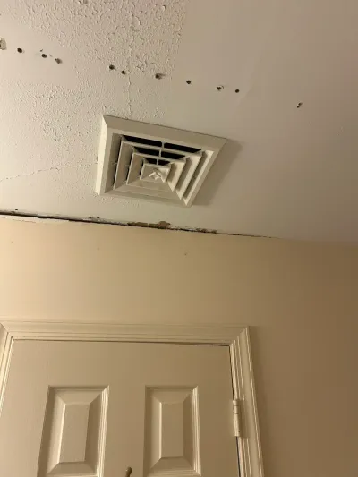 Peeling textured ceiling with square HVAC vent above a beige wall and white panel door