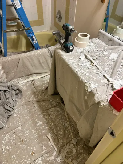 Bathroom mid-remodel with drop cloths, ladder in a yellow tub, impact driver and drywall tape on the vanity.