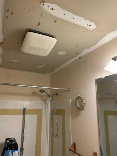 Bathroom ceiling under repair with fresh drywall patching around an exhaust fan above a tiled corner shower.