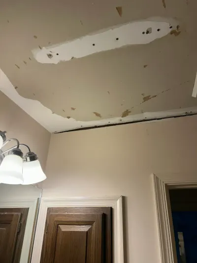 Bathroom sloped ceiling with peeling paint, a large drywall patch and open seam above an oak vanity.