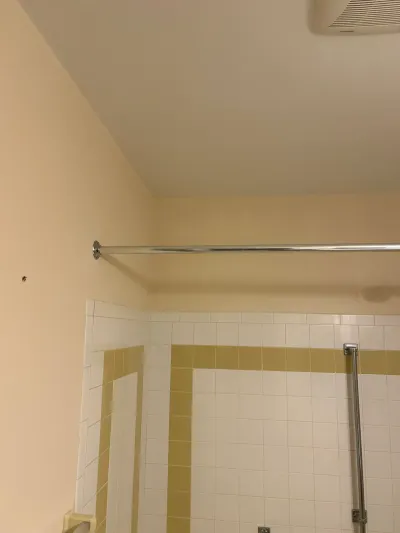 Corner of a dated bathroom showing white tile with yellow border, chrome curtain rod and ceiling exhaust fan.