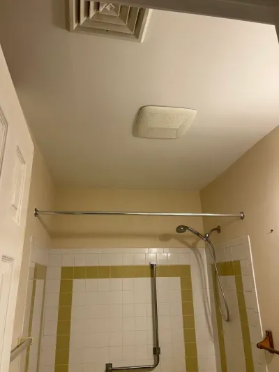 Dated bathroom shower alcove with white and gold tile, chrome curtain rod, handheld shower, and ceiling exhaust fan.