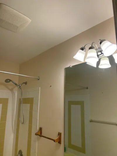 Dated bathroom corner with cream and tan tiled shower, chrome fixtures, mirror and chrome vanity light.