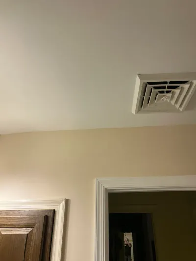White square ceiling air register above a cream wall, beside white door casing and a wood panel door.