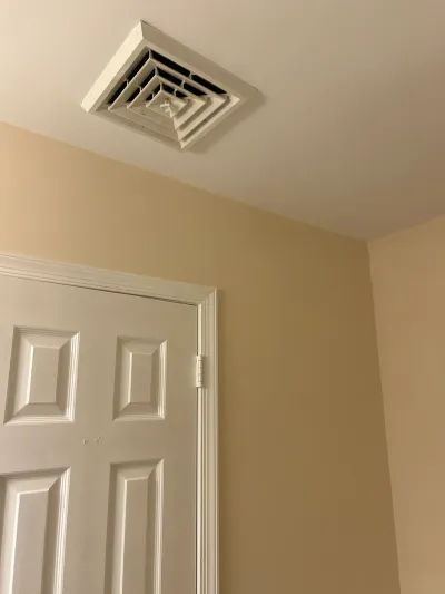 White square ceiling air vent above a beige wall and white six-panel door with painted trim.