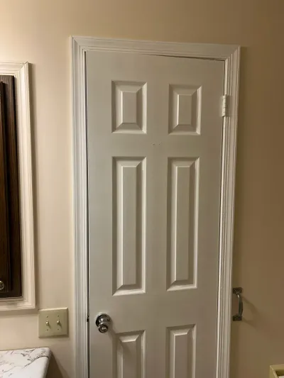 White six-panel door with chrome knob in a beige bathroom, grab bar at right and stone counter edge at left.
