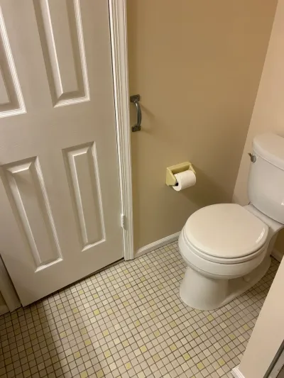 Existing bathroom corner with white toilet, tan walls and cream and yellow mosaic tile floor.