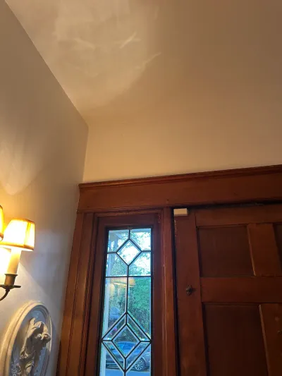 Stained wood entry door and leaded glass sidelight beneath a sloped cream ceiling, lit by a wall sconce.