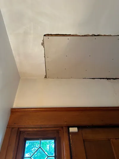 Unfinished drywall patch screwed to an upper wall below a sloped ceiling above stained wood door trim.
