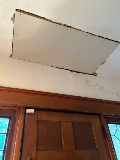 Unfinished drywall patch screwed into a ceiling above a stained wood door with water-stained plaster wall