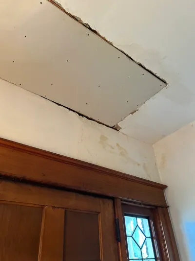 Ceiling with a freshly installed drywall patch above a stained wood door and water-stained plaster wall.