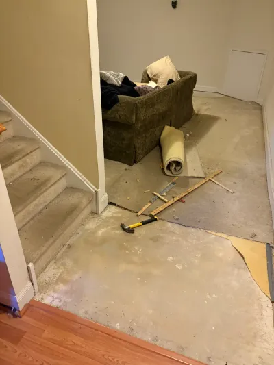 Basement mid-demolition with carpet pulled back to bare concrete, hammer and scrap wood on the floor.