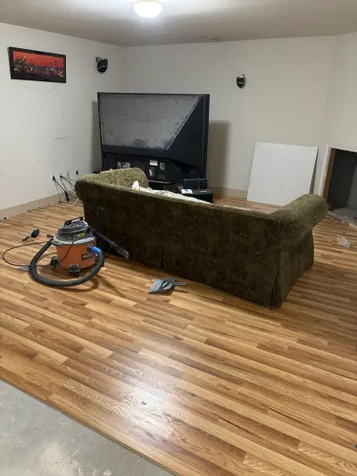 Basement room mid-remodel with new oak flooring partly installed over exposed concrete, sofa and shop vac in place.