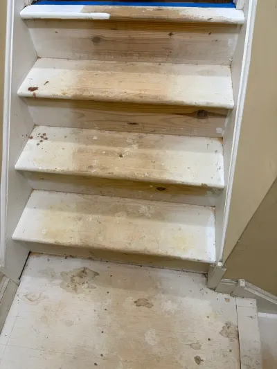 Staircase with carpet removed, exposing paint-spattered wood treads, white risers, and bare plywood landing.