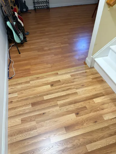 New light oak hardwood planks meeting an older amber wood floor at a seam beside a carpeted staircase.