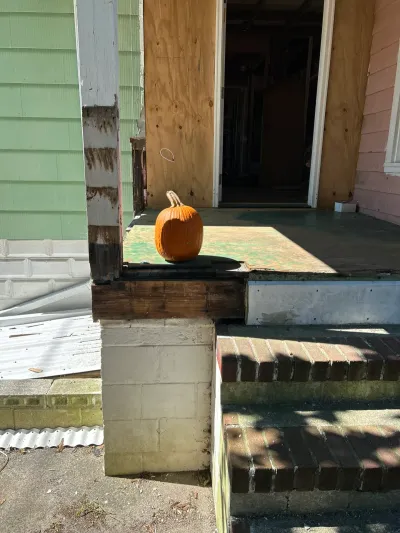 Pumpkin on the deck of a green house's front porch under renovation, doorway open and openings boarded with plywood.