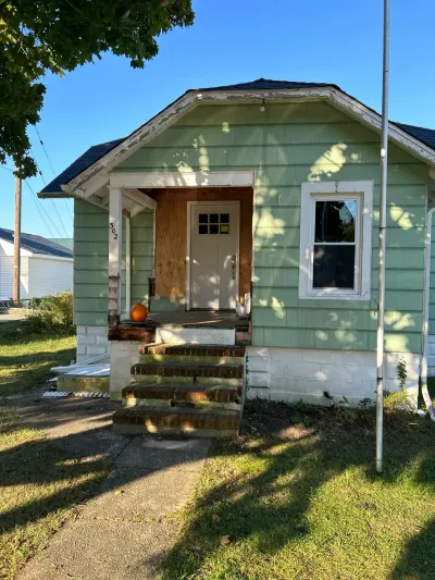 Mint green bungalow exterior mid-renovation with brick steps, white front door and bare plywood porch wall.