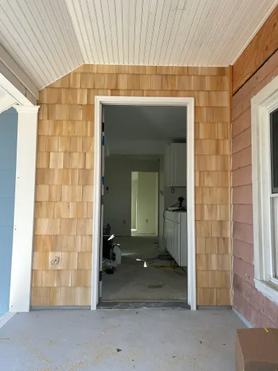 Porch doorway framed by new cedar shingles and white trim, looking into an unfinished interior room.