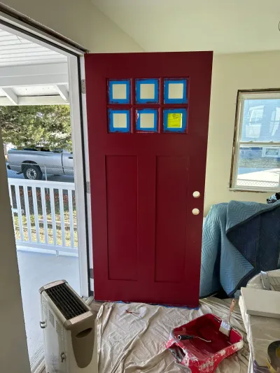 Six-lite entry door freshly painted deep red with glass masked in blue tape, drop cloth and paint tray below.