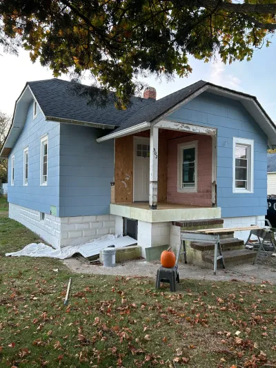 Bungalow mid-renovation with new blue-gray siding, unfinished pink porch, and tools staged on the lawn.