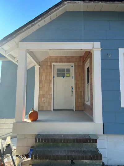 Blue-sided house entry mid-renovation with cedar shingle alcove, white columns, and brick front steps.