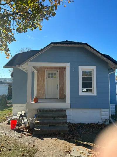Blue-sided bungalow mid-renovation with cedar shake porch wall, white trim, new front door, and brick steps.