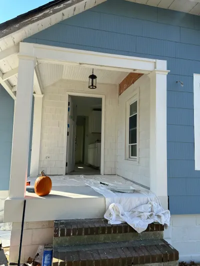 Blue-sided front porch mid-paint job with white column, beadboard ceiling, drop cloths and brick steps.