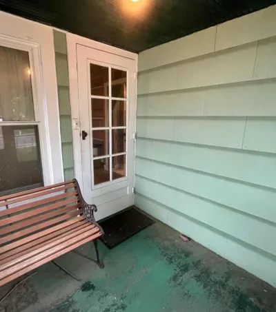 Covered porch with mint green lap siding, white glass-panel entry door, worn teal concrete floor and wood bench.