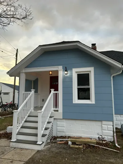 Blue lap-sided bungalow with white trim, red front door, covered porch and white railing over gray stairs.