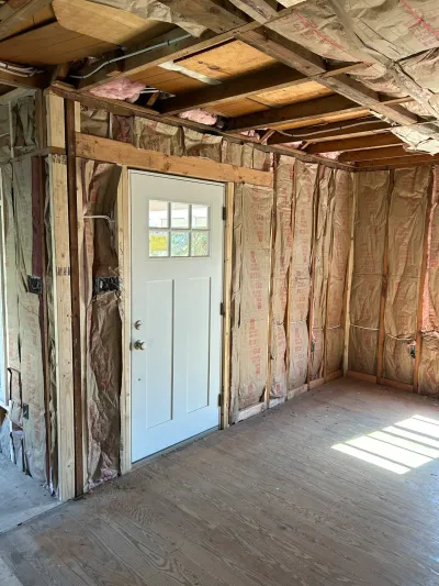 Room under renovation with exposed insulated stud walls, open ceiling joists, plywood subfloor and a white six-lite door.