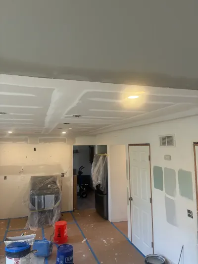Kitchen under renovation with freshly mudded drywall, plastic-wrapped appliances, paper-covered floor and paint swatches.