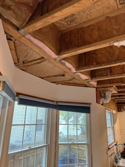 Kitchen mid-demolition with ceiling opened to exposed joists and insulation above intact bay windows.
