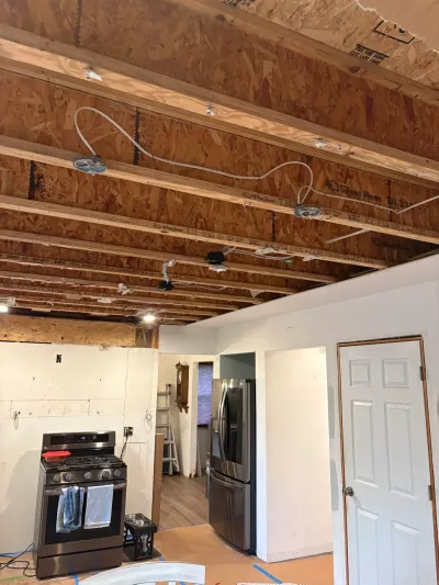 Kitchen under renovation with ceiling opened to exposed joists, loose wiring, range and refrigerator still in place.