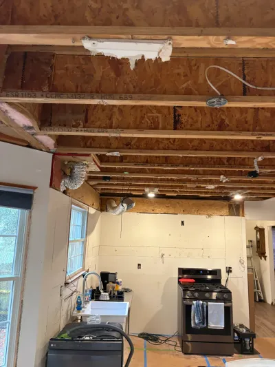 Kitchen mid-renovation with ceiling stripped to exposed joists, bare walls, temporary sink and gas range.
