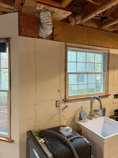 Kitchen under renovation with exposed ceiling joists, stripped walls, and a new white farmhouse sink under a window.