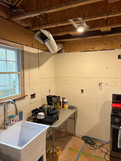 Gutted kitchen mid-renovation with exposed ceiling joists, bare walls, temporary utility sink and folding table.