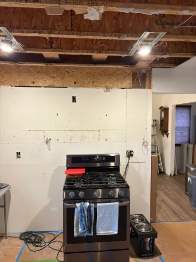 Kitchen under demolition with exposed ceiling joists, stripped walls, exposed wiring, and a stainless gas range.