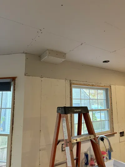 Kitchen under renovation with unfinished drywall, ceiling soffit, step ladder and chrome faucet at the window.