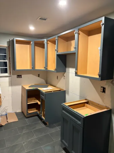 Kitchen mid-remodel with blue-gray cabinets installed, doors off, bare drywall and gray tile floor.
