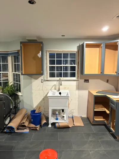 Kitchen under renovation with blue-gray cabinets, doorless boxes, temporary utility sink, and gray tile floor.